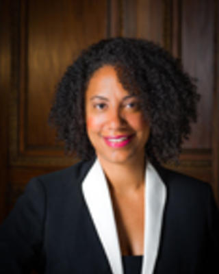 Monnique Nugent has short curly black hair. She is wearing a black blazer with white lapels.