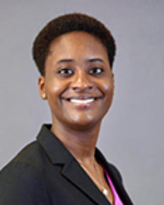 Sarah Dolisca has short coily black hair. She is wearing a pink shirt and black blazer.