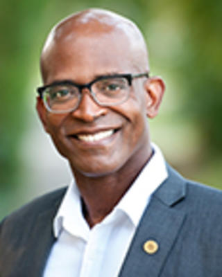 Wayne Stephens is a bald man with glasses. He is wearing a white shirt and a gray blazer with a pin.