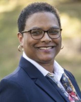 Felicia Collins is standing outside wearing a blue blazer, white shirt, and patterned scarf. She has cropped coily hair and glasses.