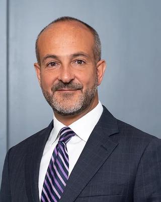 Joseph Betancourt is wearing a white shirt, blue blazer, and striped tie. He has gray facial hair.