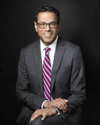 Atul Gawande has short black hair and glasses. He is wearing a gray suit with a patterned tie and sitting with his hands folded in front of him.