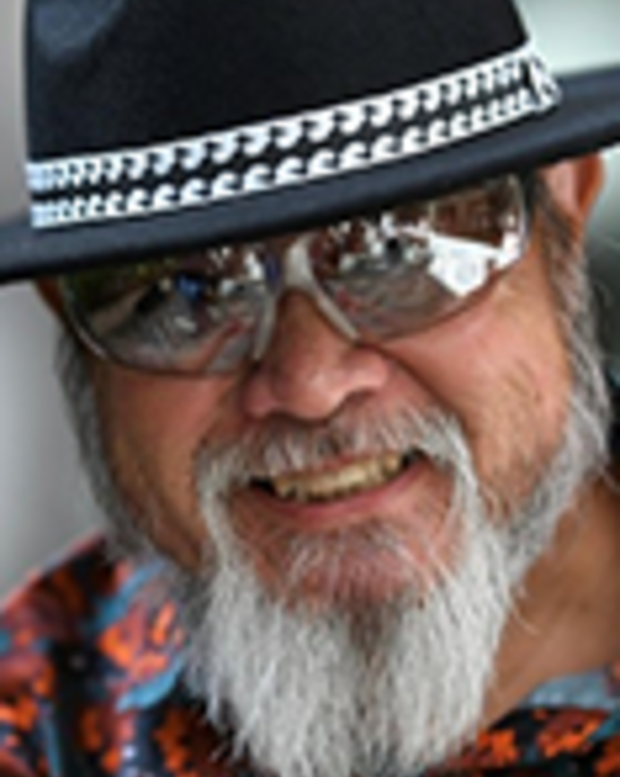 Ricardo Custodio is wearing a beaded black hat, silver sunglasses, and a printed shirt. He has a gray beard and moustache.