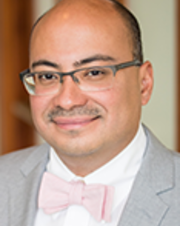 Lenny Lopez is a bald man with glasses and a moustache. He is wearing a white shirt, pink tie, and light gray blazer.