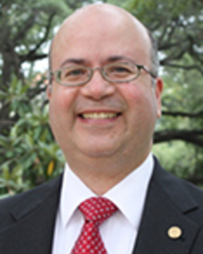Octavio Martinez is a bald man with glasses. He is standing outside in front of some greenery and wearing a suit.
