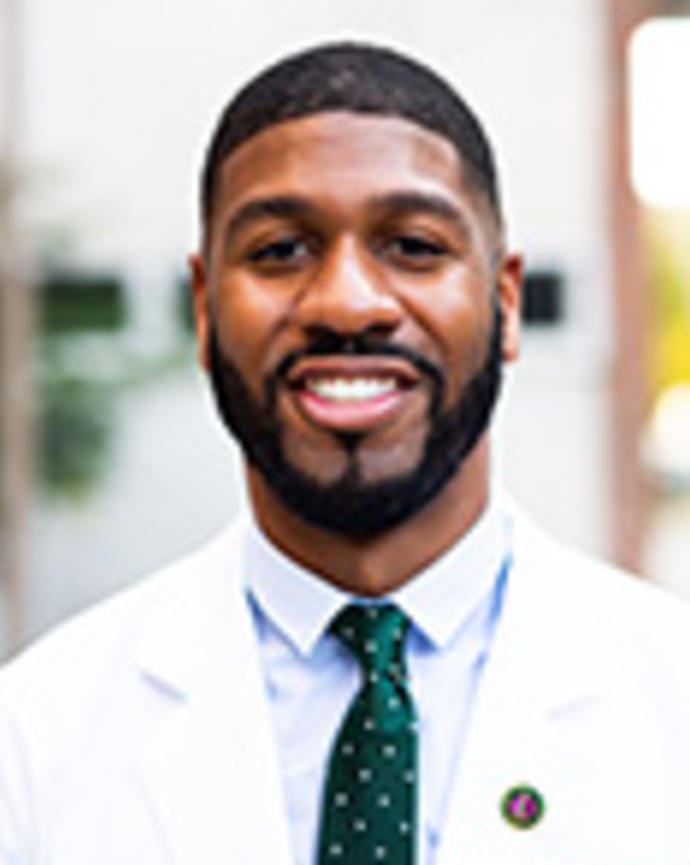 Raymond Dawkins has short black hair and facial hair. He is wearing a white coat and green patterned tie.