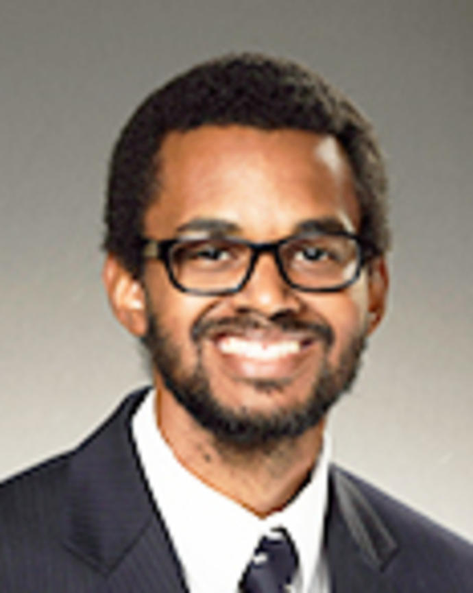 Alexander Rodgers has short coily black hair, facial hair, and glasses. He is wearing a suit.