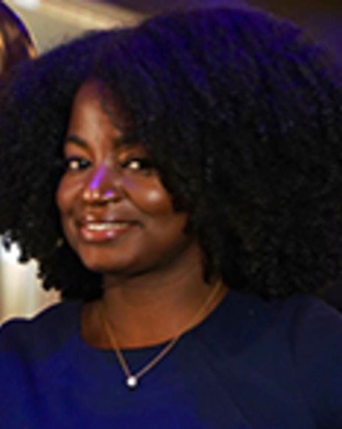 Magdala Chery has short curly black hair. She is wearing a blue dress and necklace.