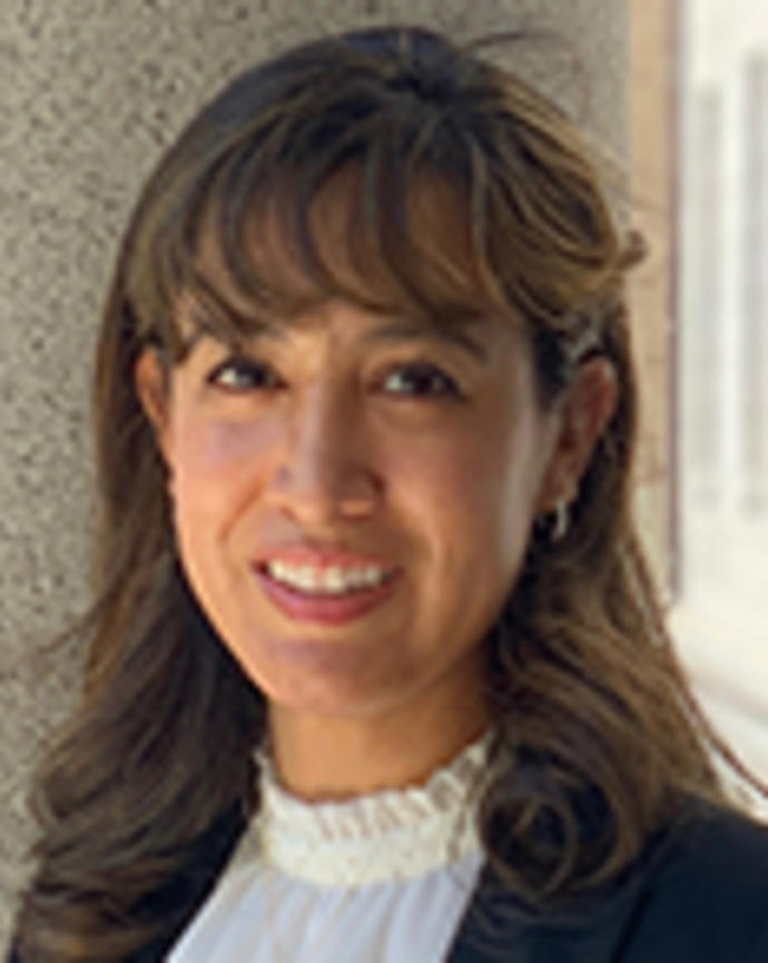 Carine Davila has medium-length brown hair with bangs. She is wearing a ruffled white blouse and black blazer.