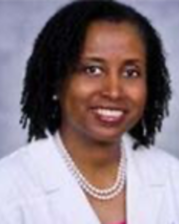 D'Nyce Williams has chin-length coily black hair. She is wearing a white coat, pink shirt, and pearl necklace.