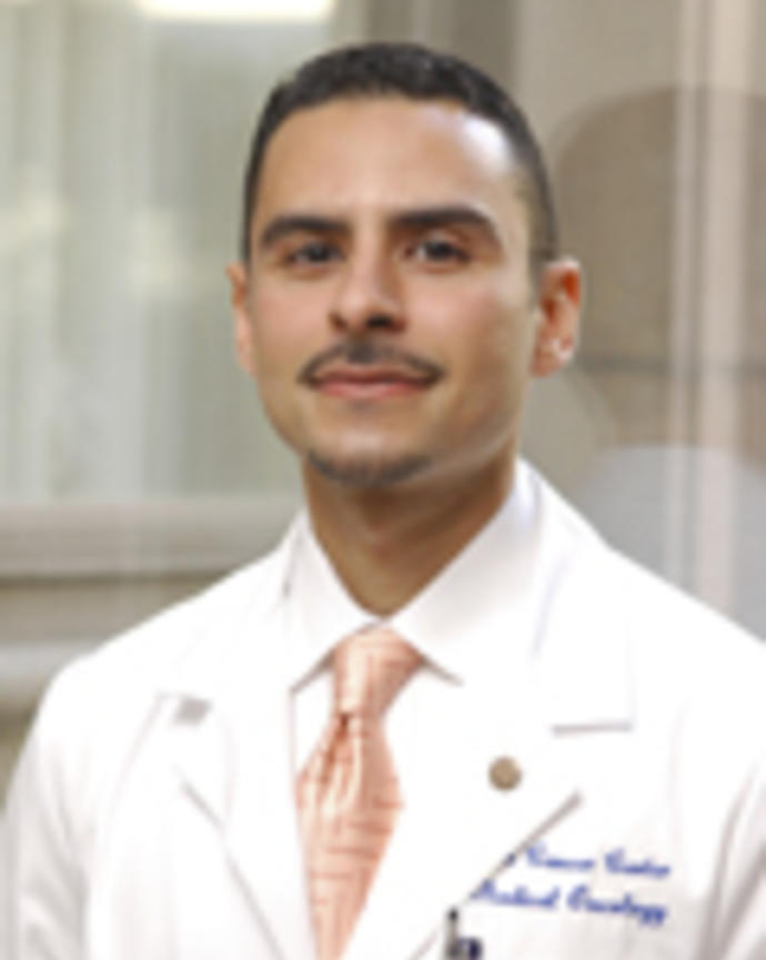 Aymen Elfiky is wearing a white coat, white shirt, and peach tie. He has short brown hair and a moustache.