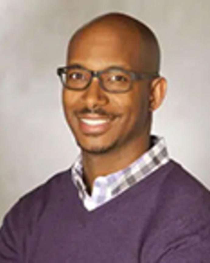 E. Elon Joffre is a bald man with glasses. He is wearing a purple plaid shirt and purple sweater.