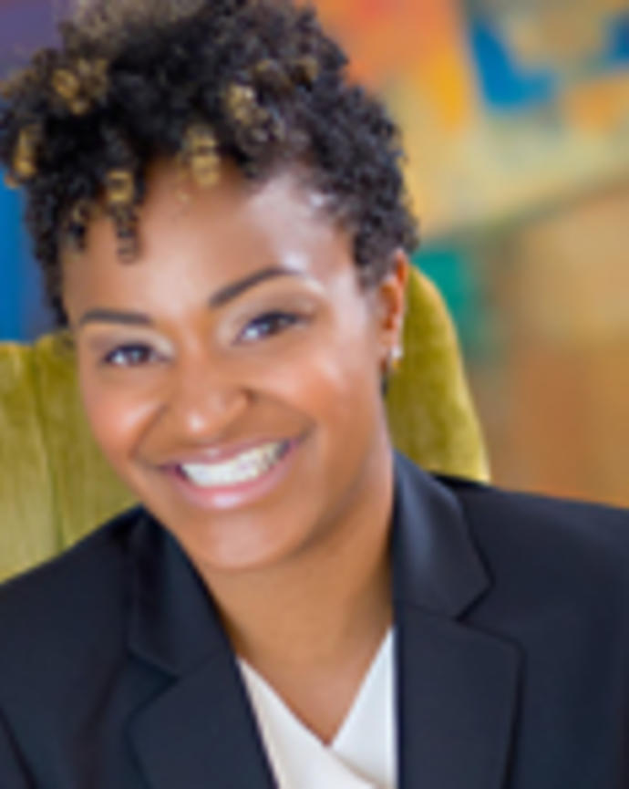 Talita Jordan has short curly black hair with blonde highlights. She is wearing a white blouse and blazer.