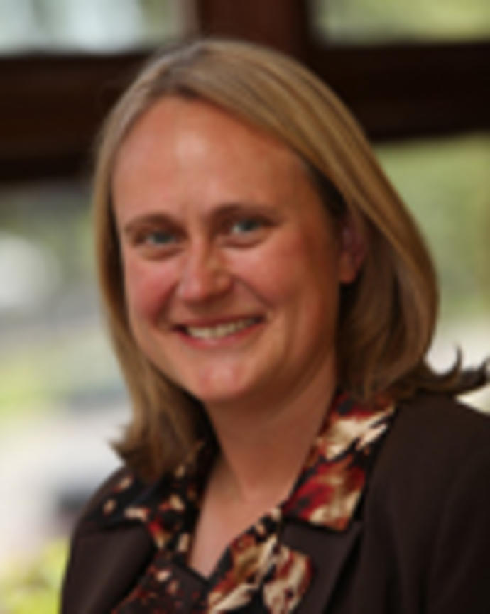 Anne Newland has medium length blonde hair. She is wearing a patterned blouse and blazer.