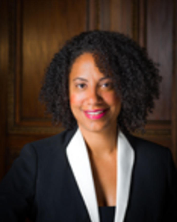 Monnique Nugent has short curly black hair. She is wearing a black blazer with white lapels.
