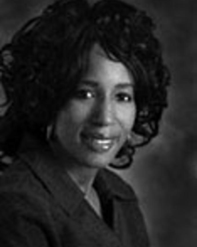 Sheila Roundtree has curly hair and is wearing a black blazer. 