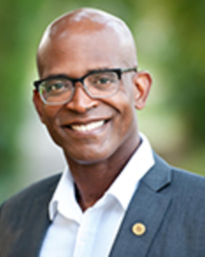 Wayne Stephens is a bald man with glasses. He is wearing a white shirt and a gray blazer with a pin.
